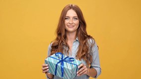 Smiling ginger woman in denim shirt holding gift and looking at camera over yellow background
