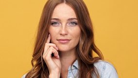 Close up of Smiling sly ginger woman in denim shirt holding arm on cheek and looking around over yellow background