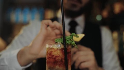 Professional bartender at fancy expensive bar in hotel, in tuxedo or suit, prepares mojito or dark and stormy cocktail made of rum and ginger beer, twists lemon peel around glass for flavor and aroma