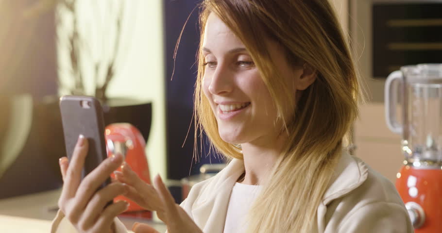 A woman in the kitchen while having breakfast and sipping tea or milk in the cup sends a message or calls with the phone and smiles. Concept of: social network, message, technology.