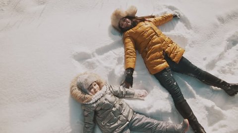 Young mother and little daughter making angels on the snow. Top view with copyspace. Family have fun in sunny winter day outdoors