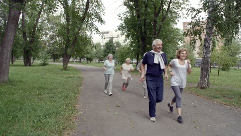 Tilt down shot of six elderly people jogging in park during group workout on warm summer day, front view