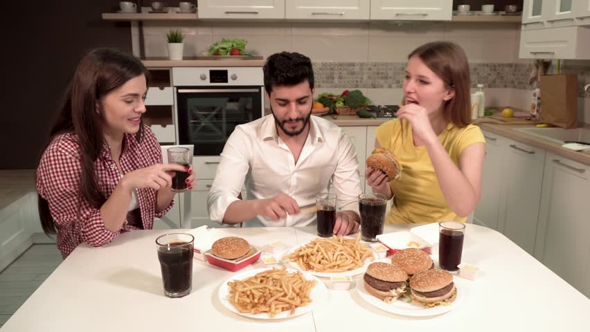 Cheerful friends having dinner, eating french fries, hamburgers and drinking cola, spending quality time in comfortable apartment