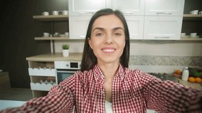 Pretty caucasian girl in red checked shirt having video chat, cheerful teen communicating via smartphone, indoor shot in light, spacious kitchen