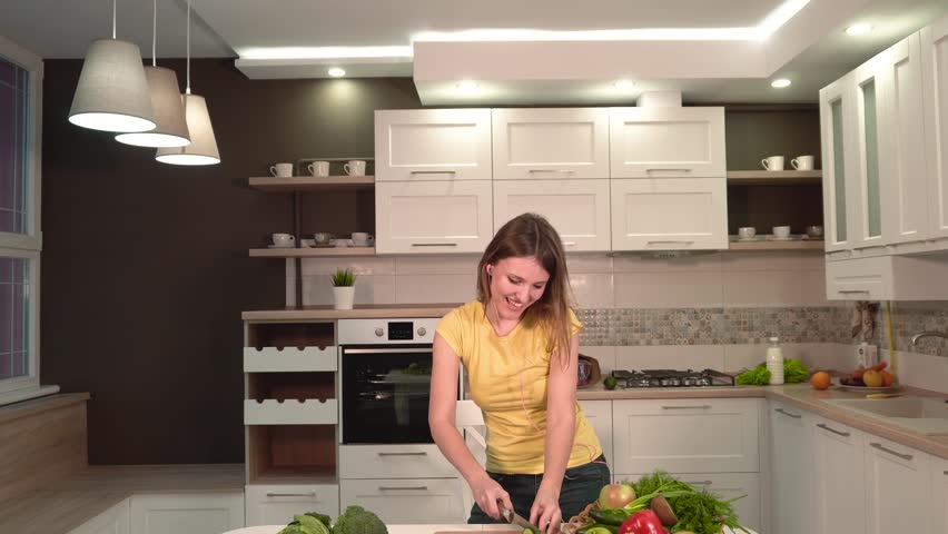 Good-looking slender girl having fun as cooking, wearing casual blue jeans and yellow t-shirt, cutting cucumber while dancing in headphones, indoor shot in large spacious kitchen