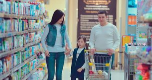 Family of three rolling a trolley with products along the supermarket, looking around and talking