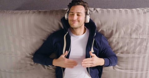 A man lying on the bed listens to music with wireless headphones and relaxes. The man dances and dreams with music. Concept of: music, relaxation, dream.