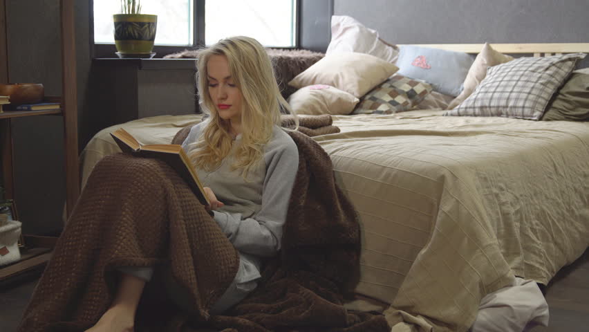 Young blonde is sitting on the floor in a blanket next to the bed and is reading a book.