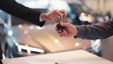 Young man giving keys of car to buyer. Men shaking hands in beautiful car dealership on background of bought cars.