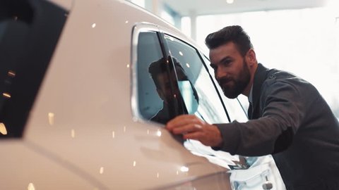 Young man with beard looking carefully at car in car dealership. Guy touching side window of beautiful shiny white car.