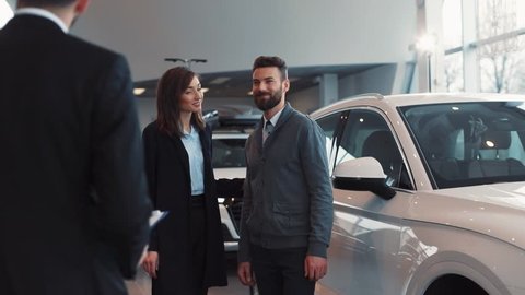 Young couple in love buying new car in car dealership. Happy people hugging. Young attractive man with beard opening door for girl. Young pretty woman sitting.