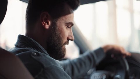 Handsome young man with beard sitting at wheel of car, talking. Guy choosing car, touching chair, wheel, viewing interior of automobile .