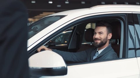 Guy in casual clothes opening car door and sitting on driver's seat. Seller giving keys. Young confident man with beard chewing gum, turning on light in car's interior.