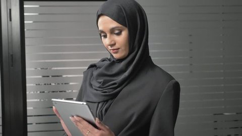 A young beautiful girl in black hijab uses a tablet, looks at photos. Arab women in the office. 60 fps