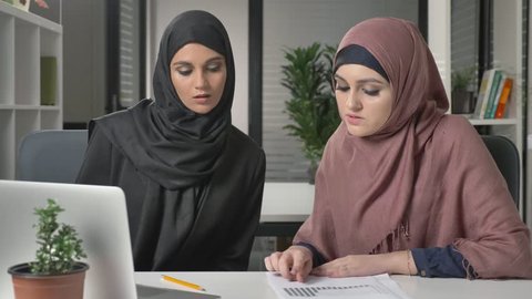 Two beautiful young girls in hijabs sit in the office and discuss schedules, business, dialogue, conversation. 60 fps