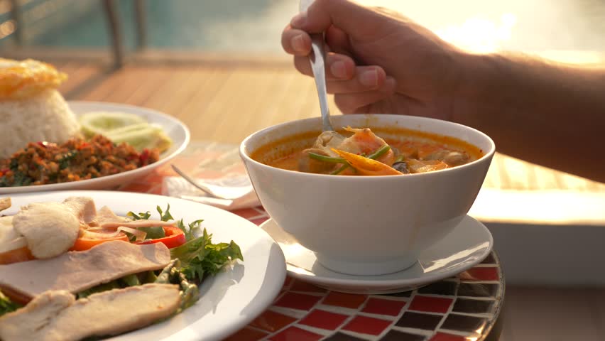 Shrimp soup is a Thai dish with a sour and spicy taste. the man is eating Tom Yam at a table by the pool. 4k, slow motion, close-up. concept of tourism tourism