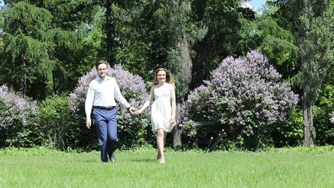 Woman and man run on grass in park with lilac at sunny day, slow motion