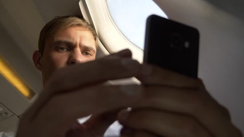 4k, close-up. Portrait of a handsome young man who looks at his smartphone while sitting by the window of an airplane during a flight.