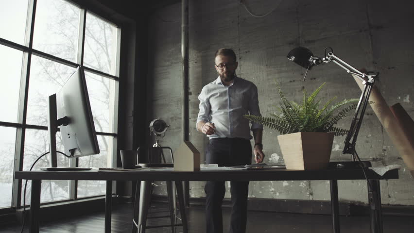 A modern male hipster works as an architect, stands in front of a table in front of a panoramic large window. Stylish workplace: office in loft style, stylish accessories on the table. Royalty-Free Stock Footage #1008114556