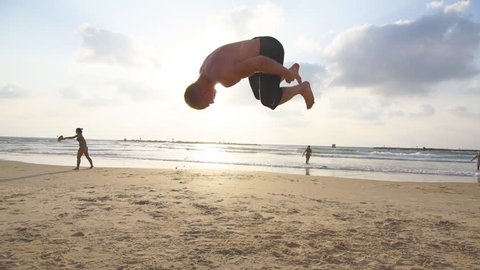 Young man doing parkour tricks on sea beach at sunset. Freerunner guy showing various jumps while running on ocean shore at sundown. Acrobatic stunts and somersaulting on sunny summer day. Slow motion