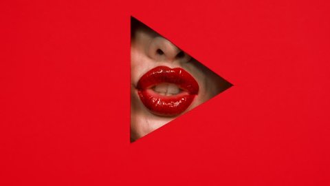 Chubby beautiful girl lips in triangular hole of paper red background.
Bright red lipstick on lips of woman. Girl shows her lips in the keyhole. Button play. Concept of advertising cosmetics.