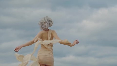 Elegant woman is wearing eccentric suit with waving fabric strips is going out from camera. Back view of her amazing slim body, picturesque clouds in background