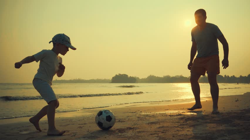 Father and son playing together with ball in football on the beach under sunset background