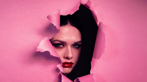 Face beautiful girl with bright make-up, with pouting lips, with red lipstick, appears in torn opening in pink paper background.
Creative advertising women's cosmetics. Girl peeks into hole.