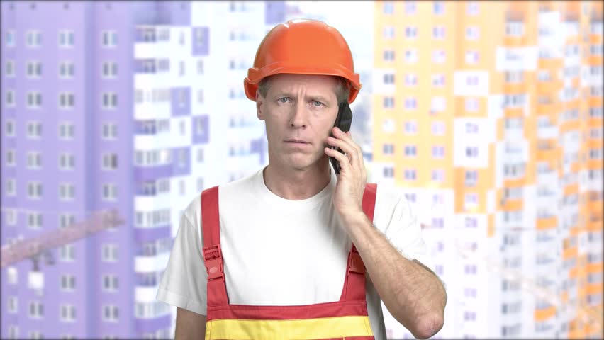 Builder with smartphone on constuctions background. Confused middle-aged engineer talking on his cell phone, blurred background.