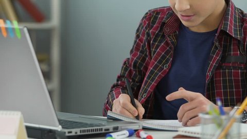 Teenager doing math homework in notebook and checking answer in laptop, close up