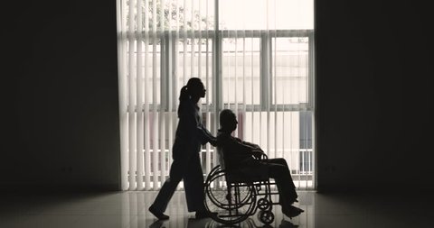 Silhouette of elderly man sitting on wheelchair and pushed by his nurse near the window at home. Shot in 4k resolution