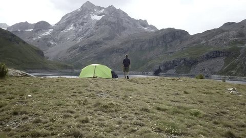 Drone shot, Man on a hike standing by its tent near a stunning mountain lake in the heart of the French Alps. Man hiker standing in nature arms outstretched. Young male arms wide open in the European