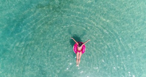 AERIAL. Top view of young pretty sexy girl swimming with pink swimming mattress at the ocean, wearing bikini and sunglasses, view on ocean, relax and having fun on vacation.