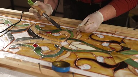 Skilled workman is processing copper tape of stained-glass panel. He is using tin solder and soldering iron, assembling and fixing beautiful hand made stained-glass