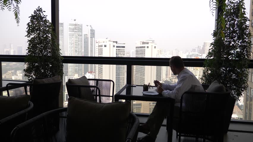 A young businessman is having breakfast in the terrace of the restaurant overlooking the skyscrapers. 4k.