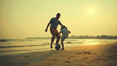 Father and son playing together with ball in football on the beach under sunset background 스톡 비디오