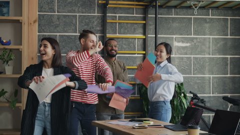 Diverse group female and male start-up business team have fun dancing party in modern office throwing paper documents and celebrating success of project