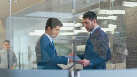 Two Businessmen Shaking Hands in the Corporate Building Hallway, They Compare Documents. Busy Office Life with ManyBusy Business Workers. Shot on RED EPIC-W 8K Helium Cinema Camera.
