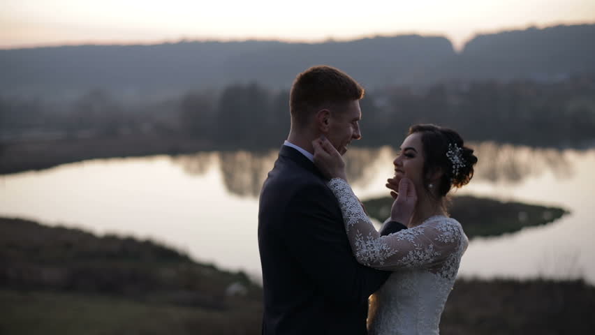 A young and beautiful wedding couple standing together near lake and making a kiss. Slow motion