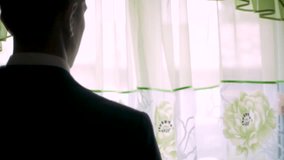Back view on man in a jacket stands at the window, his hand raised. Clip. Rear view of a man standing in front of a window and looking out it in a bedroom at home
