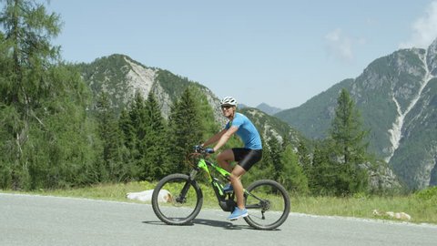 SLOW MOTION CLOSE UP: Cheerful young cyclist riding electric bike easily up a mountain road on sunny summer day. Guy cycling with ease on his e-bike. Fit male rider effortlessly ascending steep road