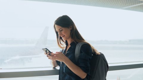 Young girl using smartphone near airport window. Happy European woman with backpack uses mobile app in terminal. 4K.