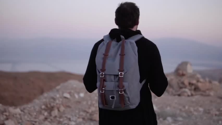 Man with backpack walking in dusk desert wilderness. Slow motion. Male wanders alone looking at scenic sunset sky.