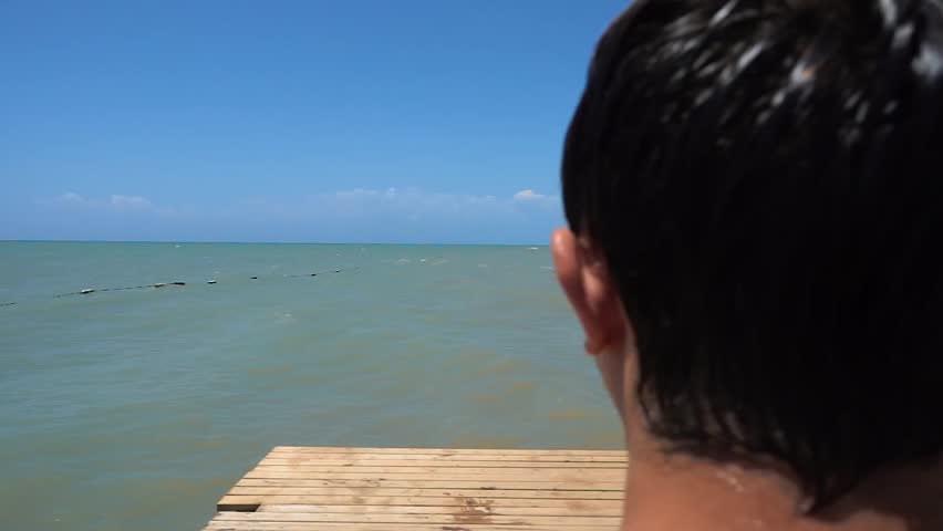 Young man runs away and during jump from pier to sea shows like. Extreme. Sunny weather. Rest at hotel. Resort. Slow motion.