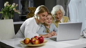 Cute little girl sitting with mom and grandmother at table, smiling and looking at laptop screen while browsing the Net