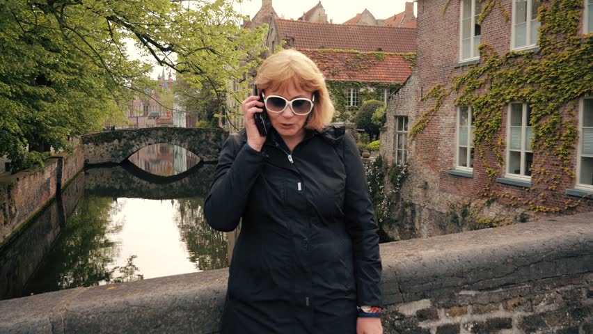 Mature businesswoman in black business coat talking on phone on street near the river canal of old stone city 4k