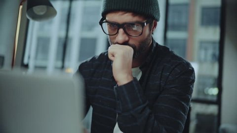 Bearded hipster man wearing eye glasses and using devices at office. Confident busy male freelancer in trendy eyewear using laptop while surfing net searching information for presentation