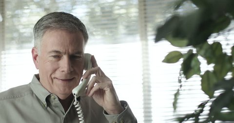 A mature Caucasian male standing in front of a sunlit window using a landline telephone engaged happily in lively conversation 4k