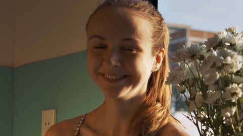 closeup pretty blonde confused girl with ponytail smiles and smells white hrizantemas flowers on windowsill