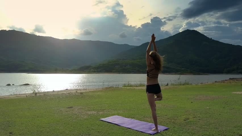 backside view slim blond girl stands in yoga pose Vrikshasana on mat near blinking under sunlight lake